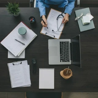 A top-down view of a doctor's desk with a person in blue scrubs writing on a patient chart. A stethoscope, laptop, paperwork, and a potted plant are also visible